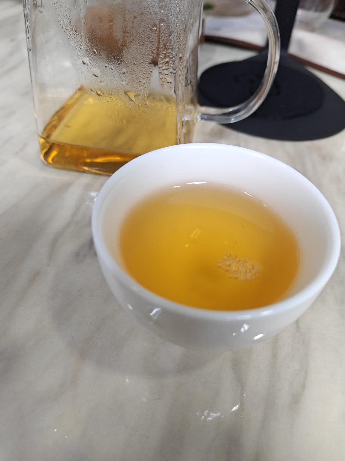 Golden fifth infusion of 2021 Jingmai raw Pu’er in a white cup, with a glass pitcher behind it.
